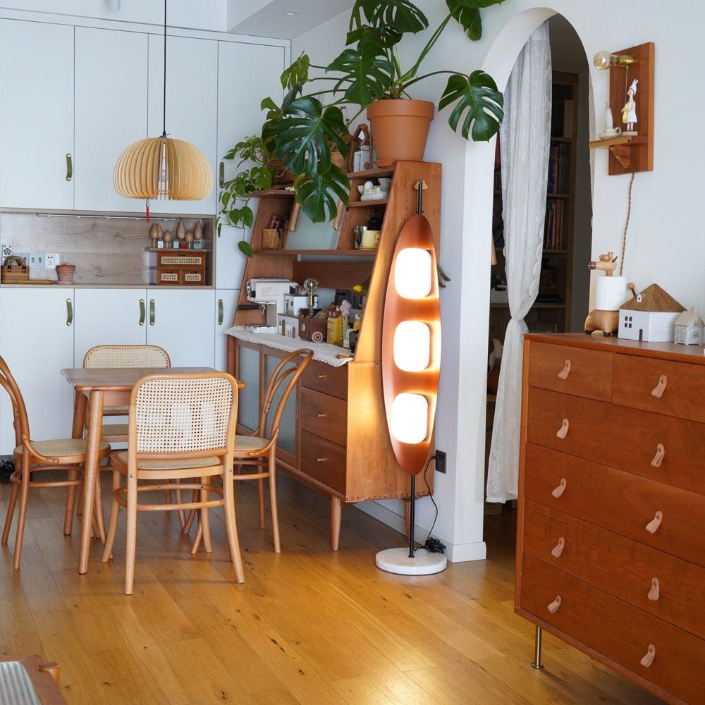Clérachic teak floor lamp standing near an archway in a wood-cabinetry kitchen and dining nook, its shades glowing warm white.