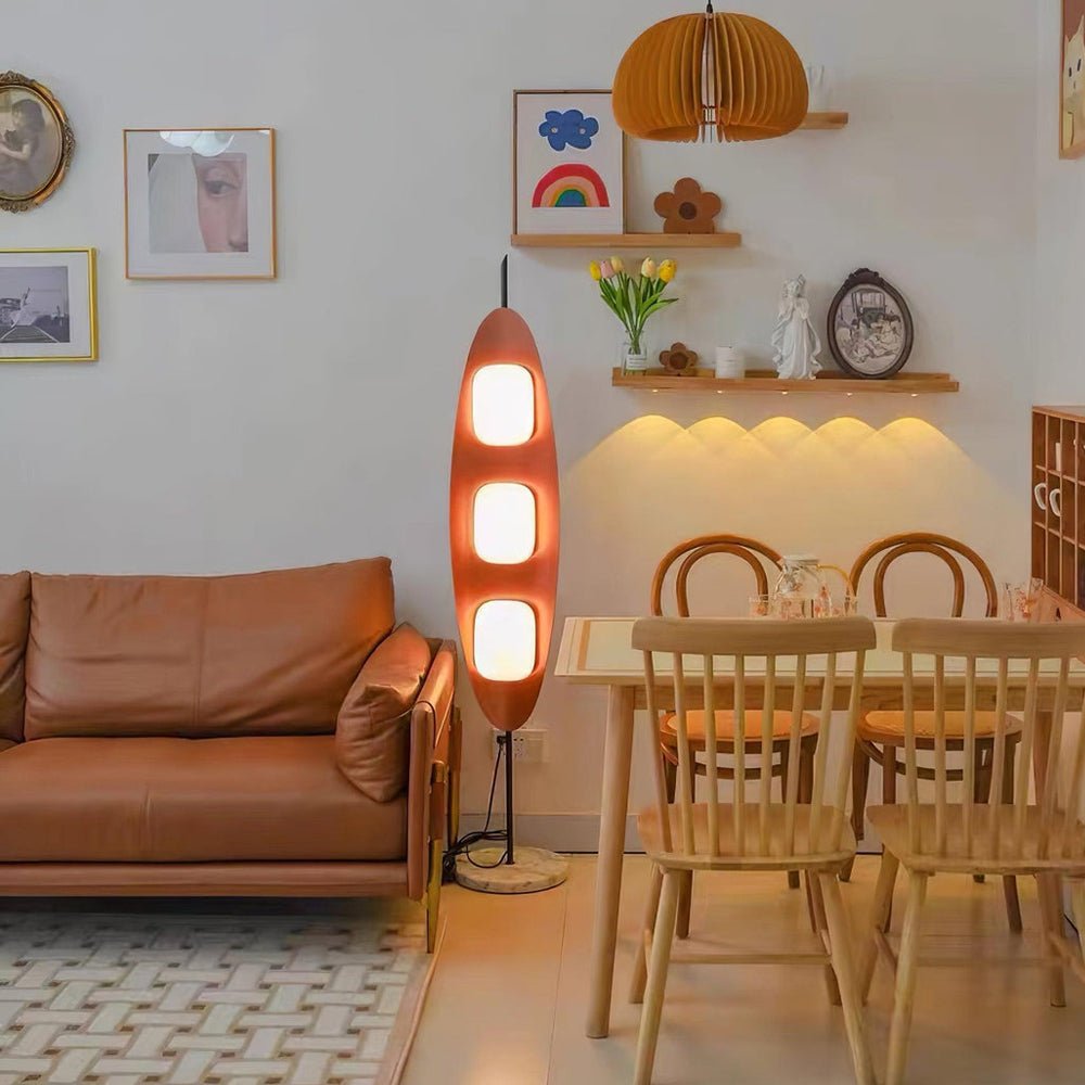 Clérachic teak floor lamp lit beside a tan leather sofa and a wooden dining table in an open-plan living and dining room.