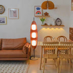 Clérachic teak floor lamp lit beside a tan leather sofa and a wooden dining table in an open-plan living and dining room.