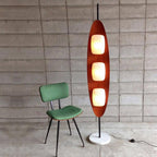 Clérachic teak floor lamp beside a green vintage chair against a bare concrete block wall, its power cord visible at the base.