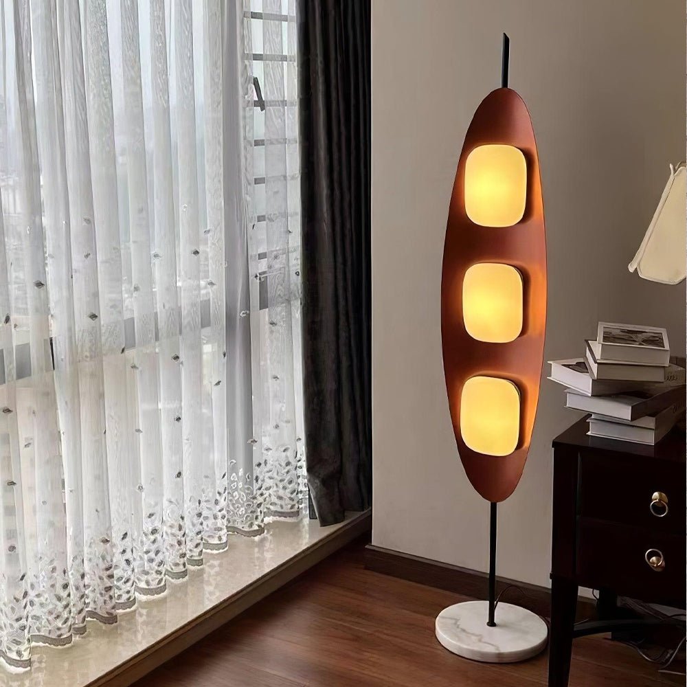 Clérachic teak floor lamp glowing beside a sheer-curtained window and a dark wood side table stacked with books at dusk.