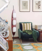 Clérachic teak floor lamp beside a striped green armchair and framed line-art prints in a colourfully rugged room.