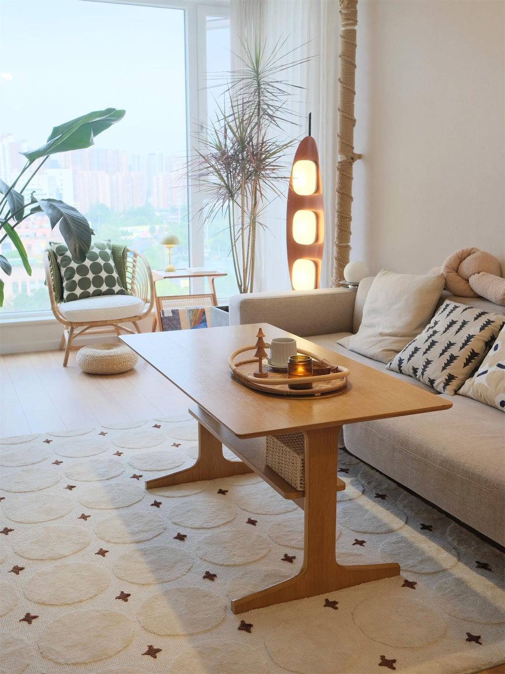 Clérachic teak floor lamp lit beside a wicker chair and coffee table, with a city skyline visible through the window.
