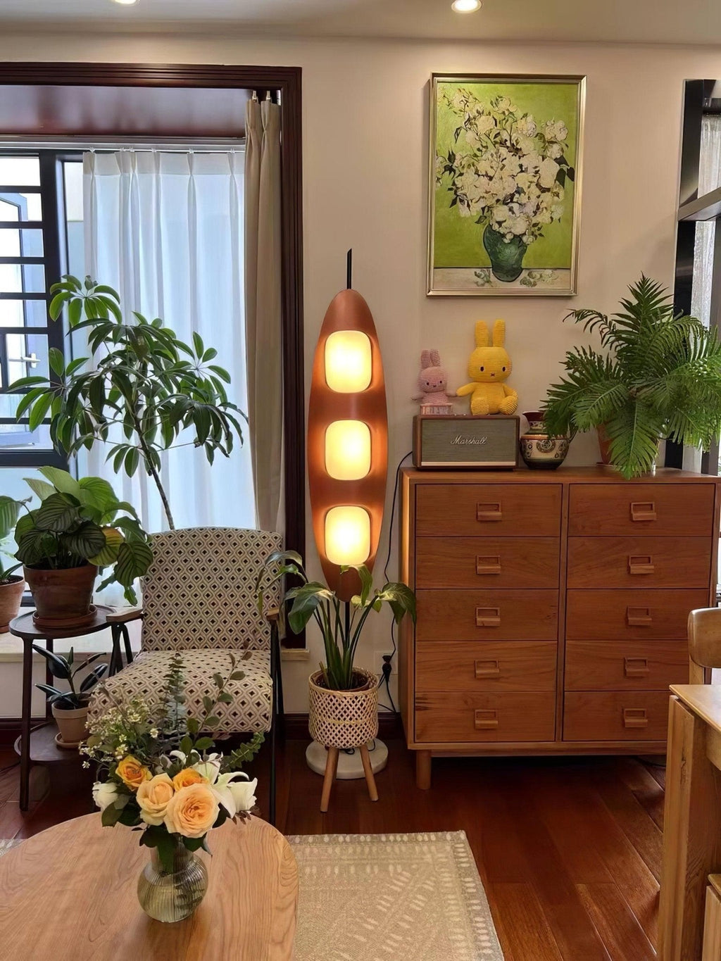 Clérachic teak floor lamp glowing beside a wooden dresser topped with flowers and framed art in a warm-toned living room.