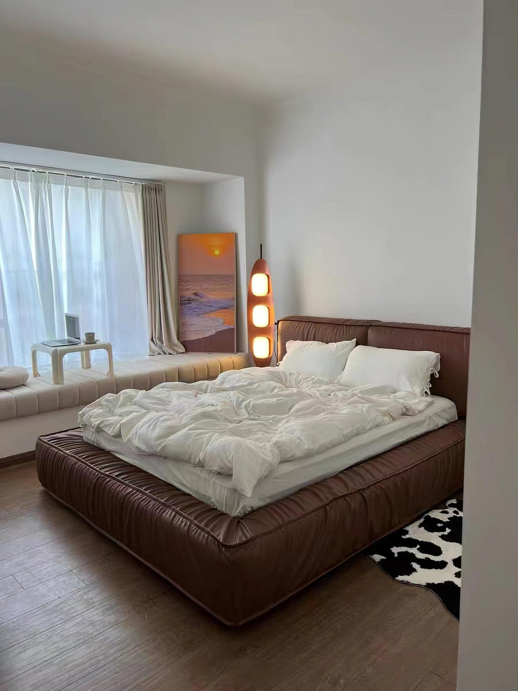 Clérachic teak floor lamp glowing beside a brown leather bed frame with white bedding and a cowhide rug on the floor.