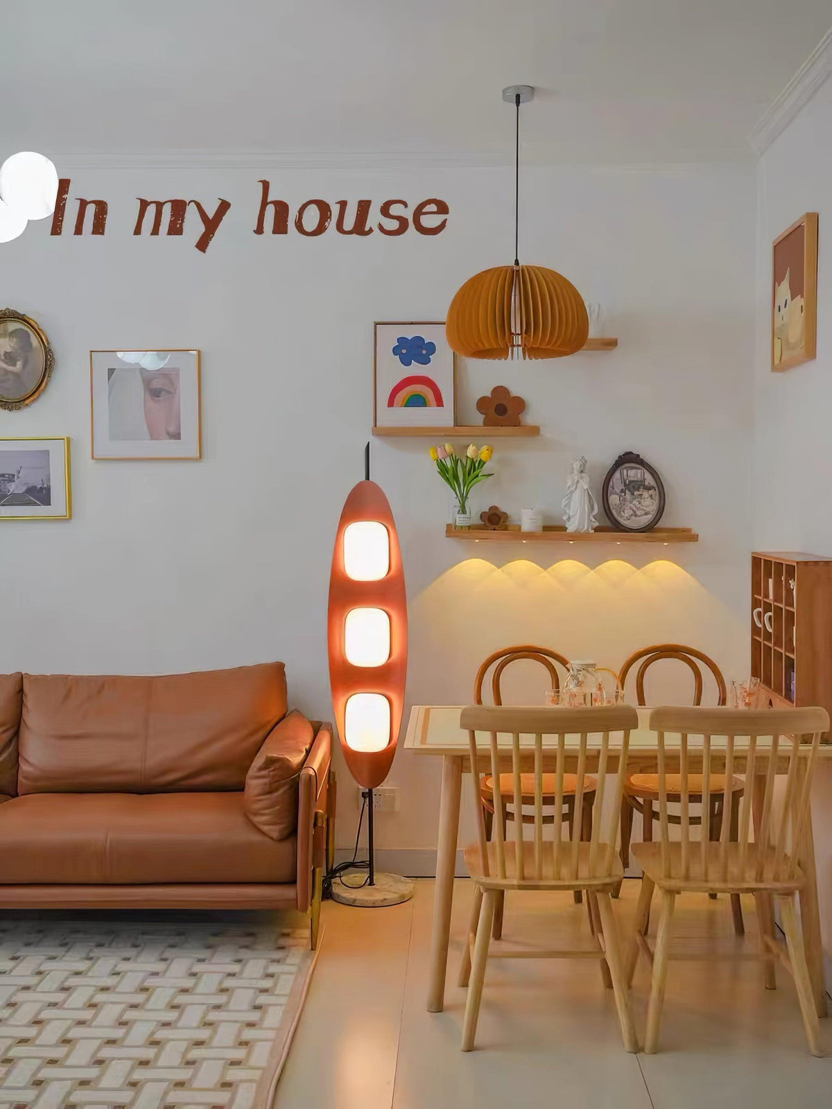 Clérachic teak floor lamp lit beside a leather sofa and wooden dining set, with a folded paper pendant on a shelf nearby.