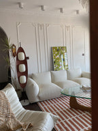 Clérachic teak floor lamp lit beside a white boucle sofa and a glass coffee table, against a wall with decorative panel moulding.