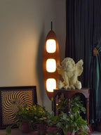 Clérachic teak floor lamp glowing in a dim corner beside a dark curtain, its light catching a small cherub statue nearby.