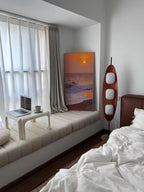Clérachic teak floor lamp lit beside a window seat with white bedding, near a small framed sunset-beach painting.