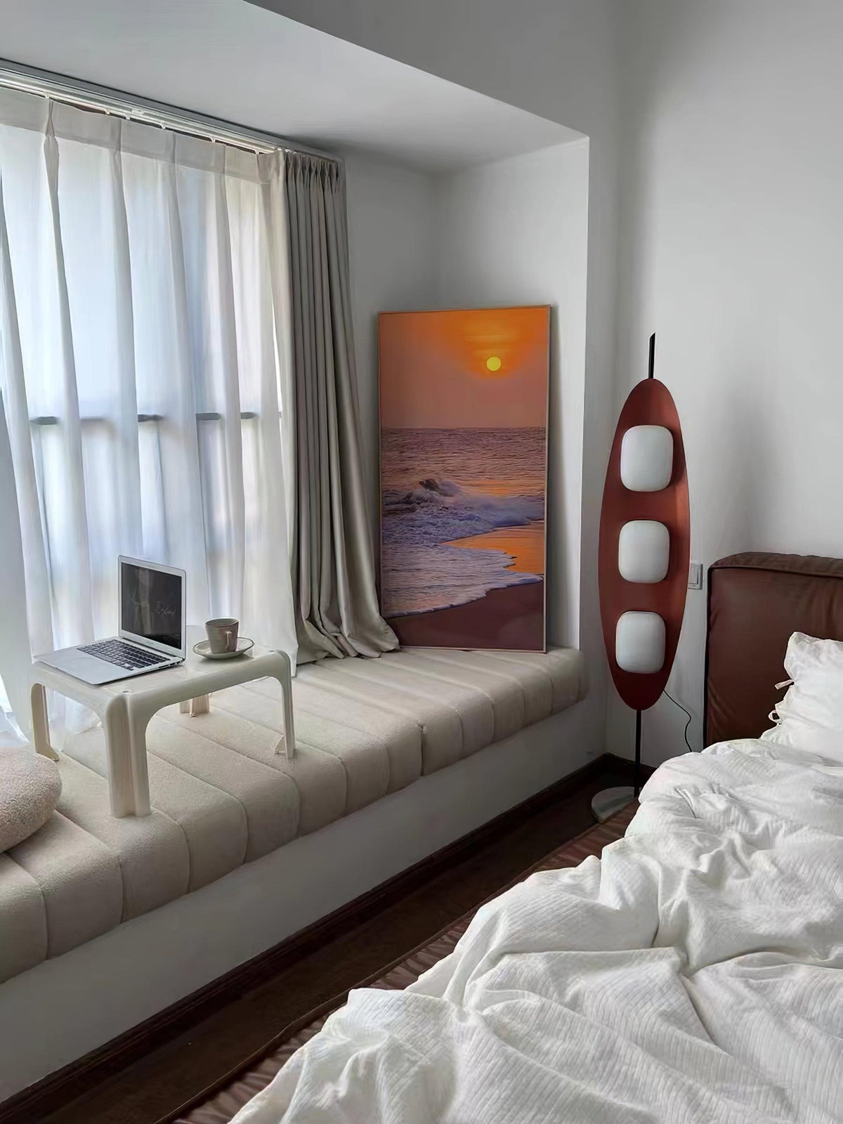 Clérachic teak floor lamp lit beside a window seat with white bedding, near a small framed sunset-beach painting.