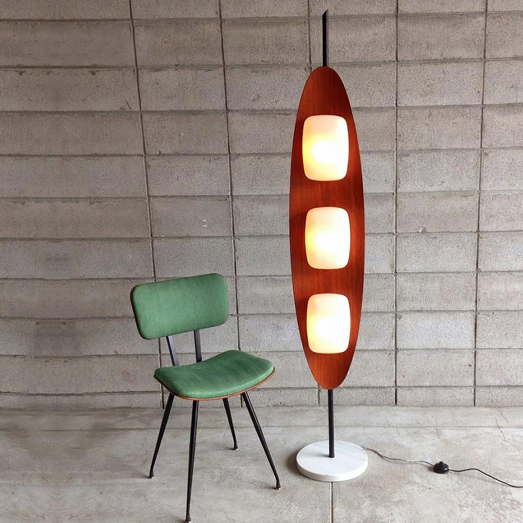 Clérachic teak floor lamp positioned beside a green velvet accent chair, set against a bare concrete block wall.