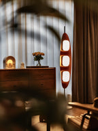 Clérachic teak floor lamp glowing beside a wood dresser and a vase of flowers, soft curtain-shadow patterns falling across it.