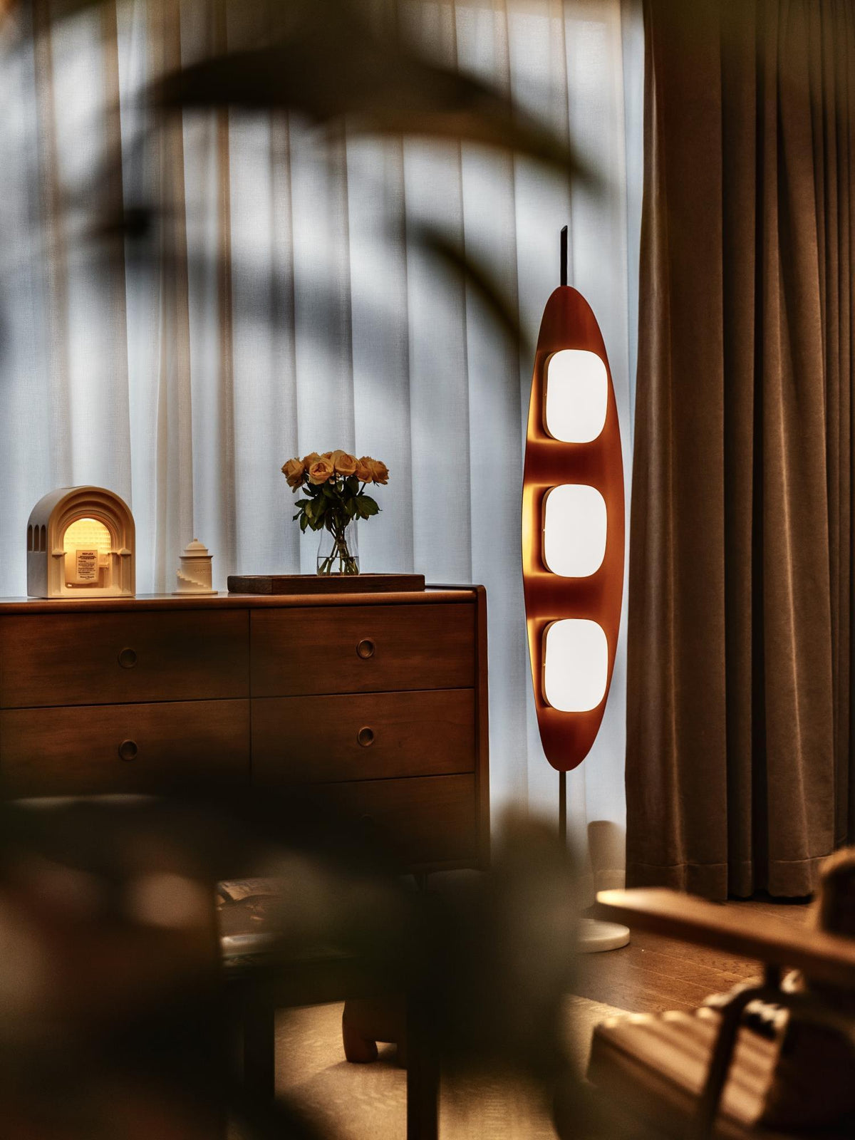 Clérachic teak floor lamp glowing beside a wood dresser and a vase of flowers, soft curtain-shadow patterns falling across it.