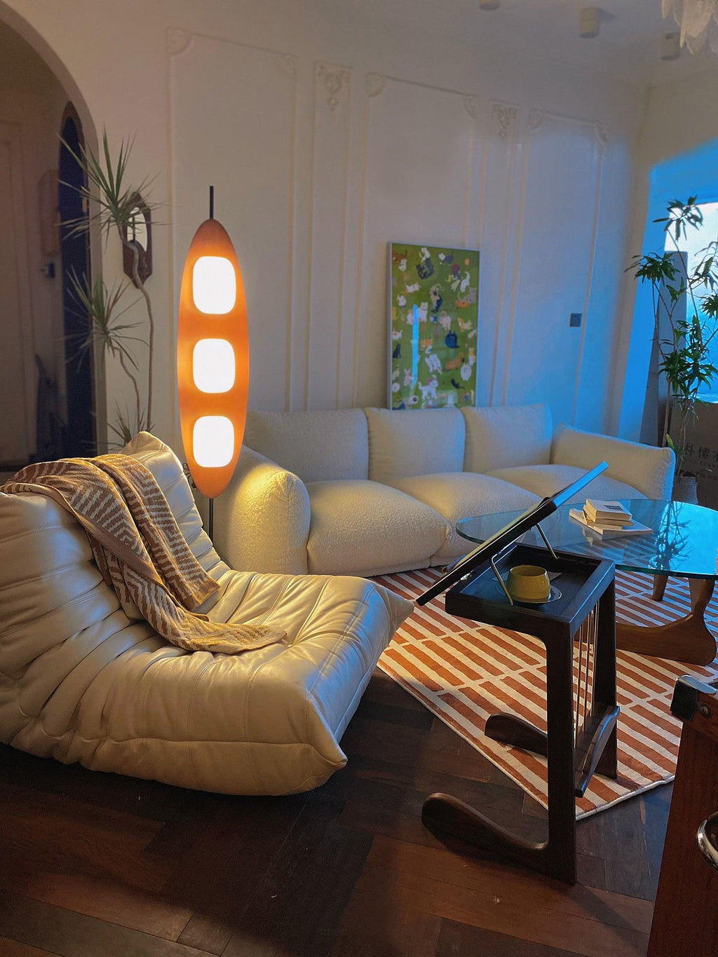 Clérachic teak floor lamp lit beside a white sectional sofa and a glass side table, the room cast in cool evening light.