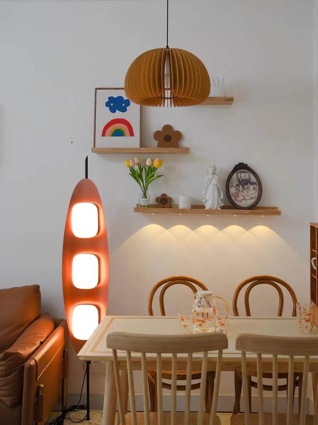 Clérachic teak floor lamp glowing near a wooden dining table, beneath a floating shelf and a folded paper pendant light.