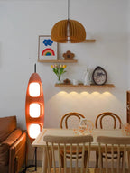 Clérachic teak floor lamp glowing near a wooden dining table, beneath a floating shelf and a folded paper pendant light.