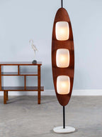Clérachic teak floor lamp shown full length on a plain grey backdrop, beside a console table with a slender bird sculpture.