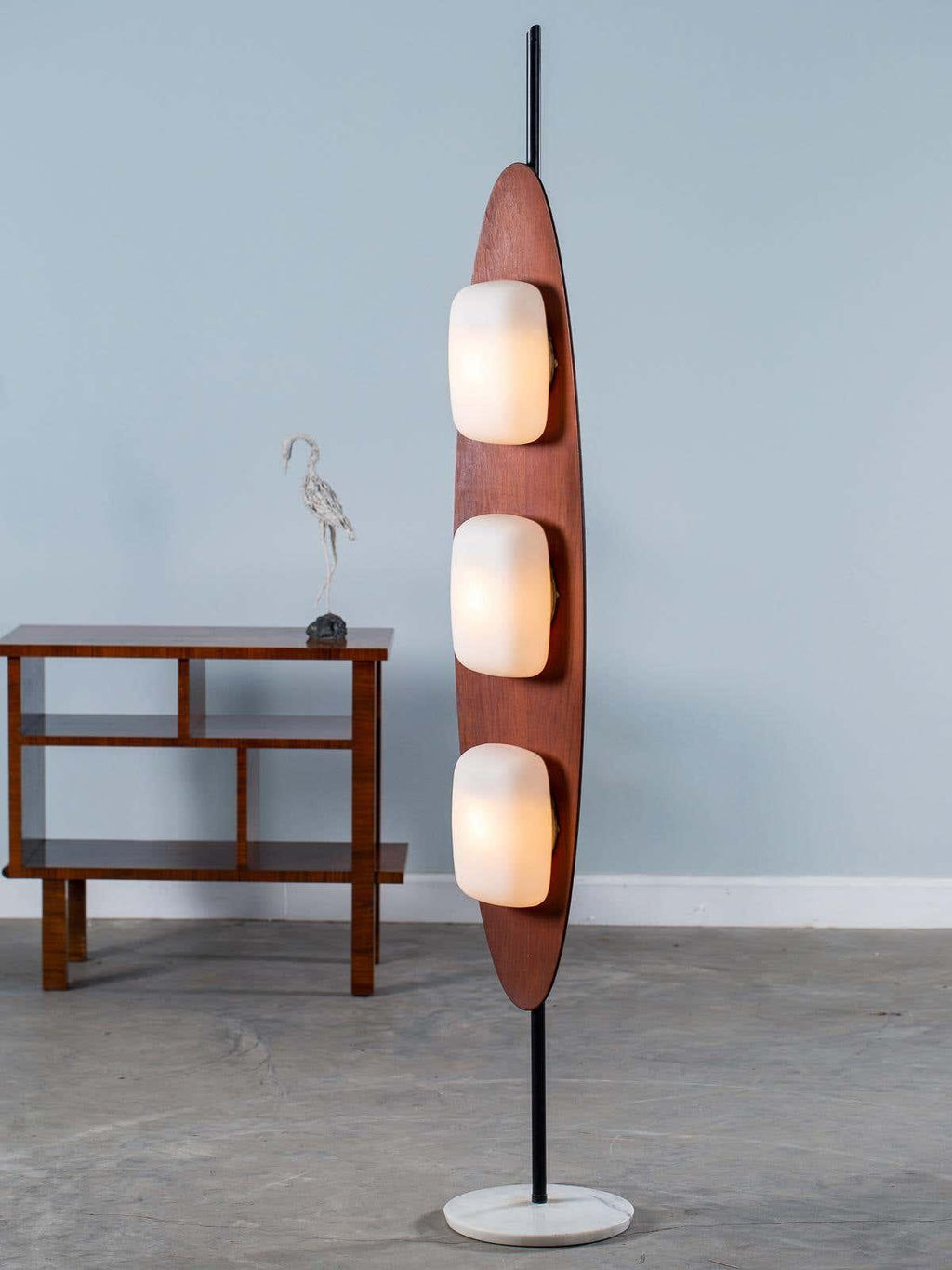 Clérachic teak floor lamp on its round marble base, photographed against a plain grey backdrop from a slightly different angle.