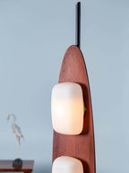 Close-up of a Clérachic teak floor lamp's lower glass shade, the carved wood body filling the frame against a grey wall.
