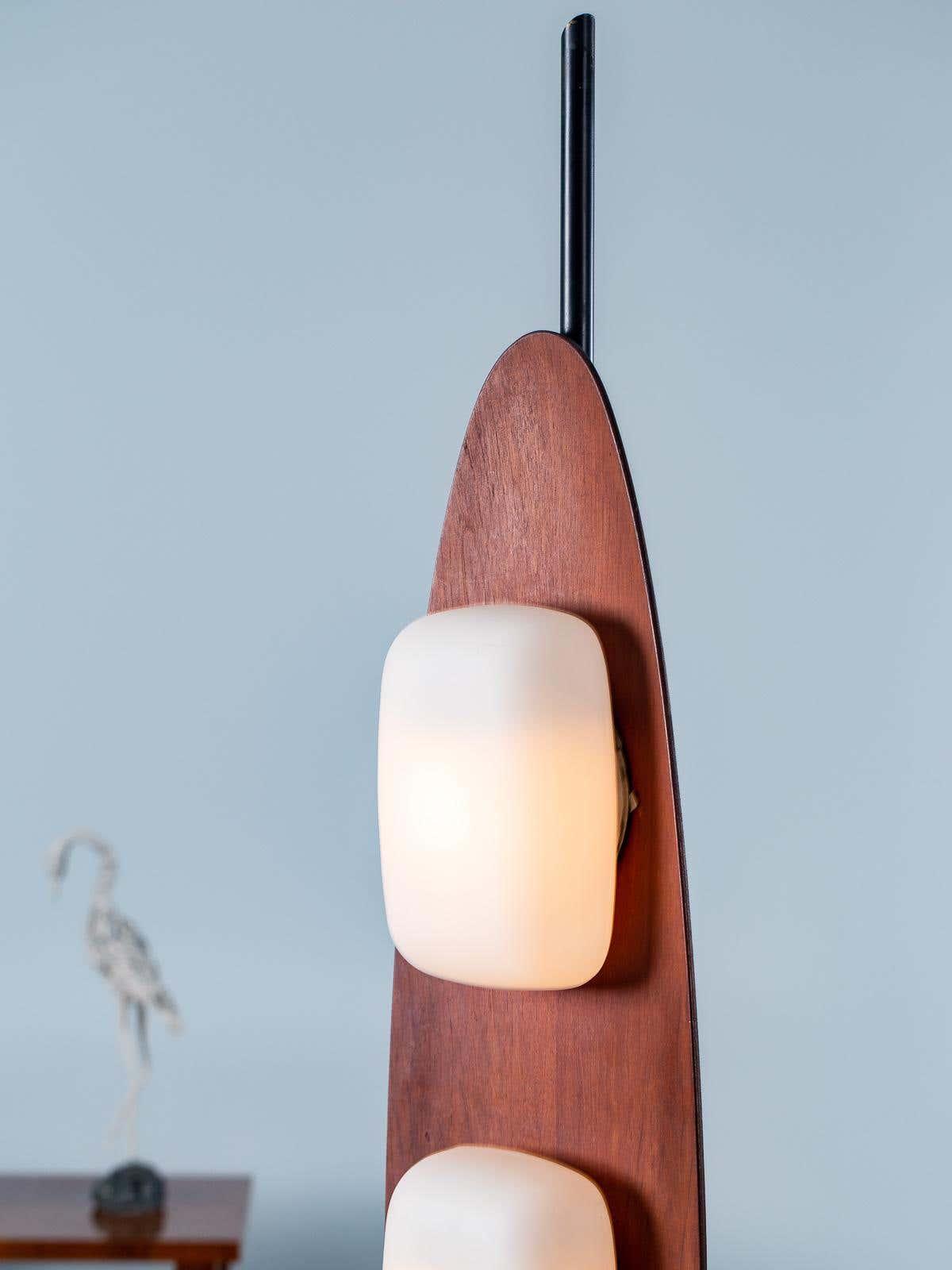 Close-up of a Clérachic teak floor lamp's lower glass shade, the carved wood body filling the frame against a grey wall.