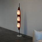 Clérachic teak floor lamp glowing near a pale sofa on dark polished flooring, evening light showing through the window behind it.