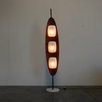 Clérachic teak floor lamp beside a wooden dining table, with houseplants and a city skyline visible through the window behind.