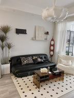 Clérachic teak floor lamp beside a black leather sofa with animal-print cushions and a checked area rug in a living room.