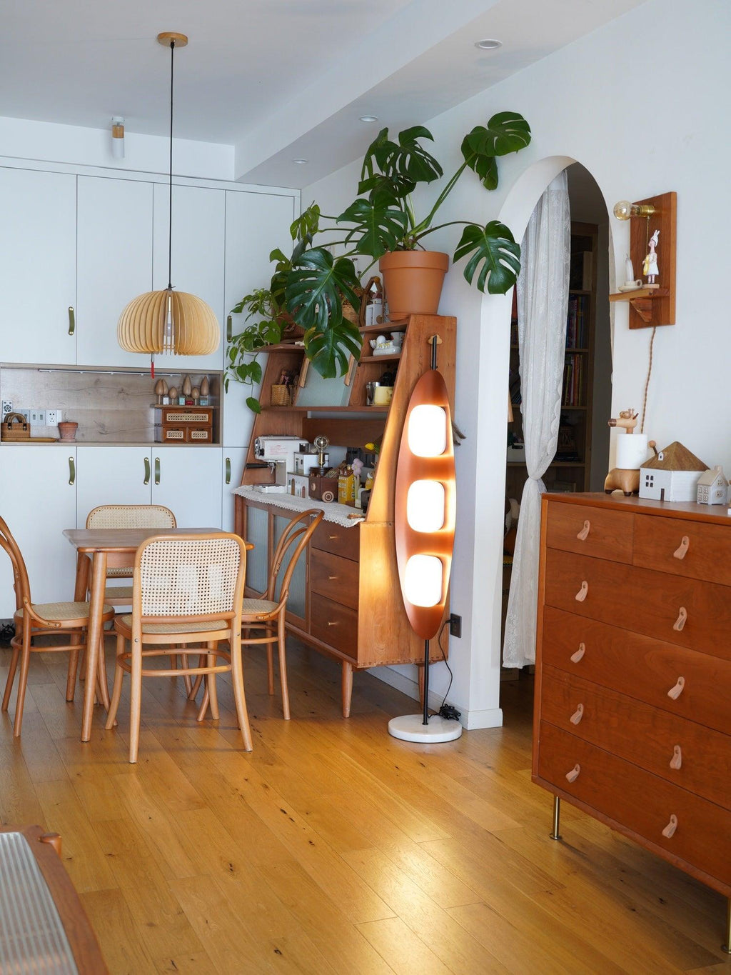 Clérachic teak floor lamp lit near a woven pendant light in a wood-cabinetry kitchen, beside a dining table and houseplant.