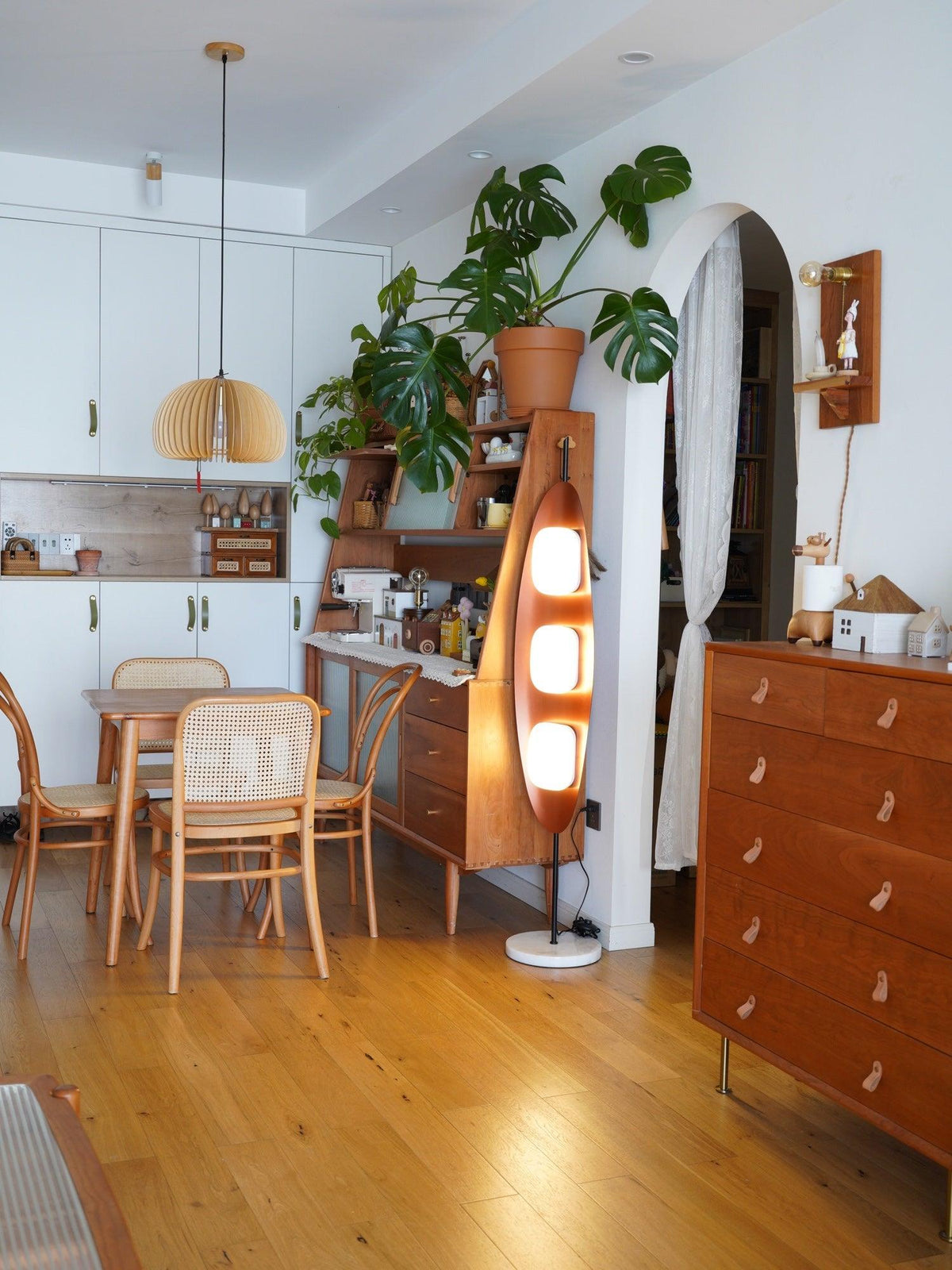 Clérachic teak floor lamp lit near a woven pendant light in a wood-cabinetry kitchen, beside a dining table and houseplant.