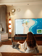 Clérachic teak floor lamp glowing beside a television, its warm light reaching a person petting a grey cat on the floor.