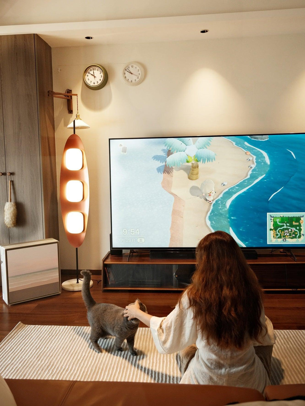 Clérachic teak floor lamp glowing beside a television, its warm light reaching a person petting a grey cat on the floor.