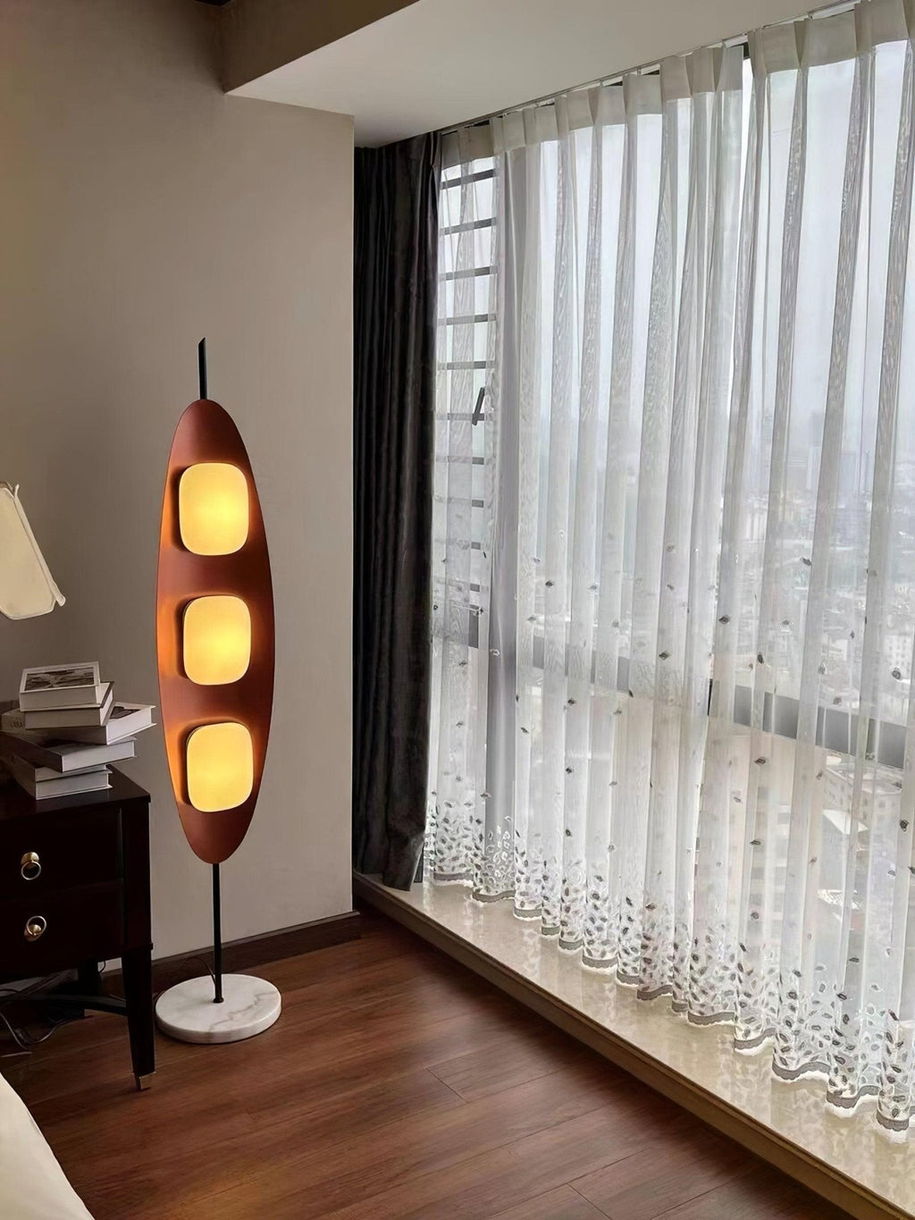 Clérachic teak floor lamp beside a sheer-curtained window and dark wood dresser drawers, lit warm against the evening light.