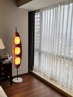 Clérachic teak floor lamp beside a sheer-curtained window and dark wood dresser drawers, lit warm against the evening light.