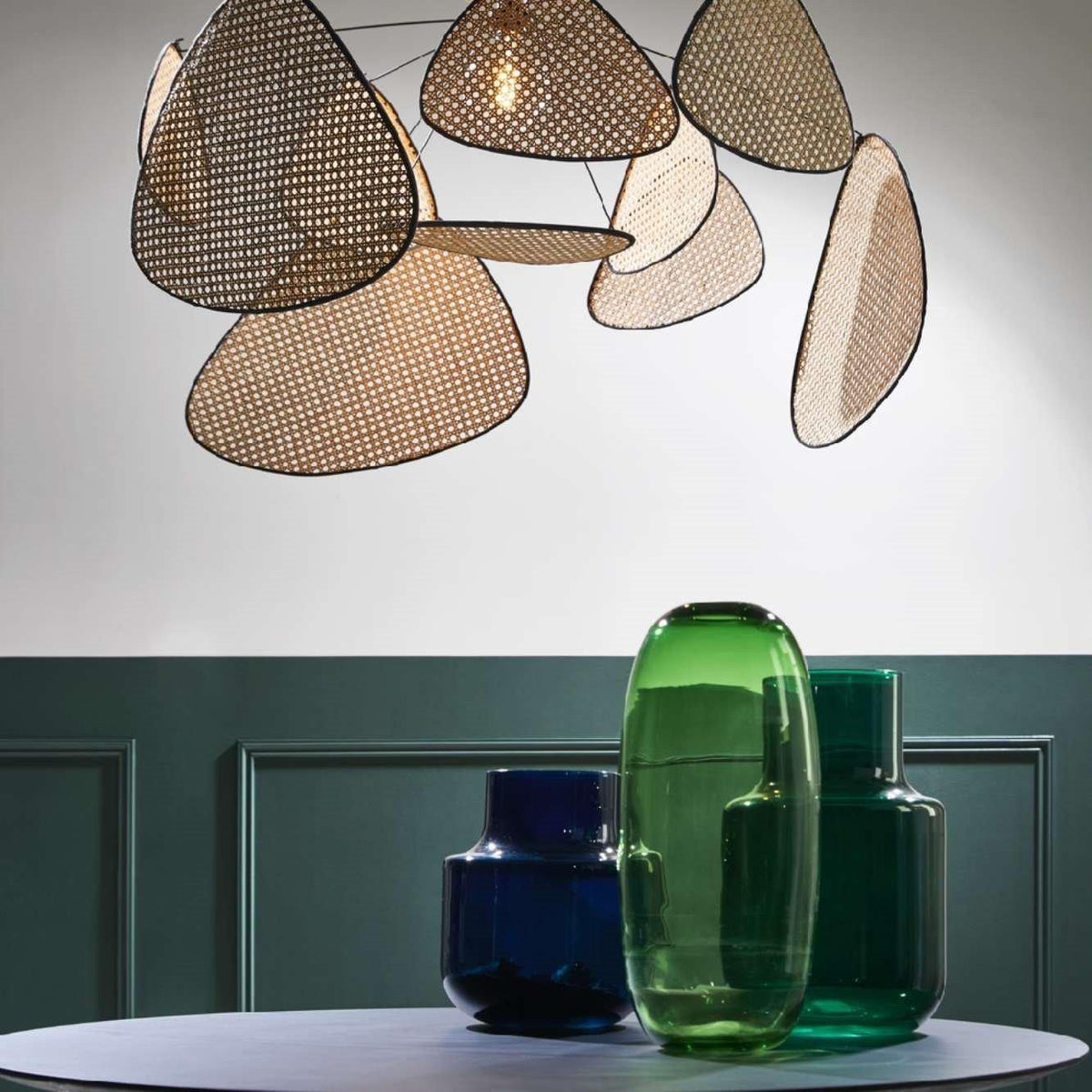A close view shows this Clérachic pendant's overlapping bamboo-weave paddles styled above coloured glass vases against a green wall.