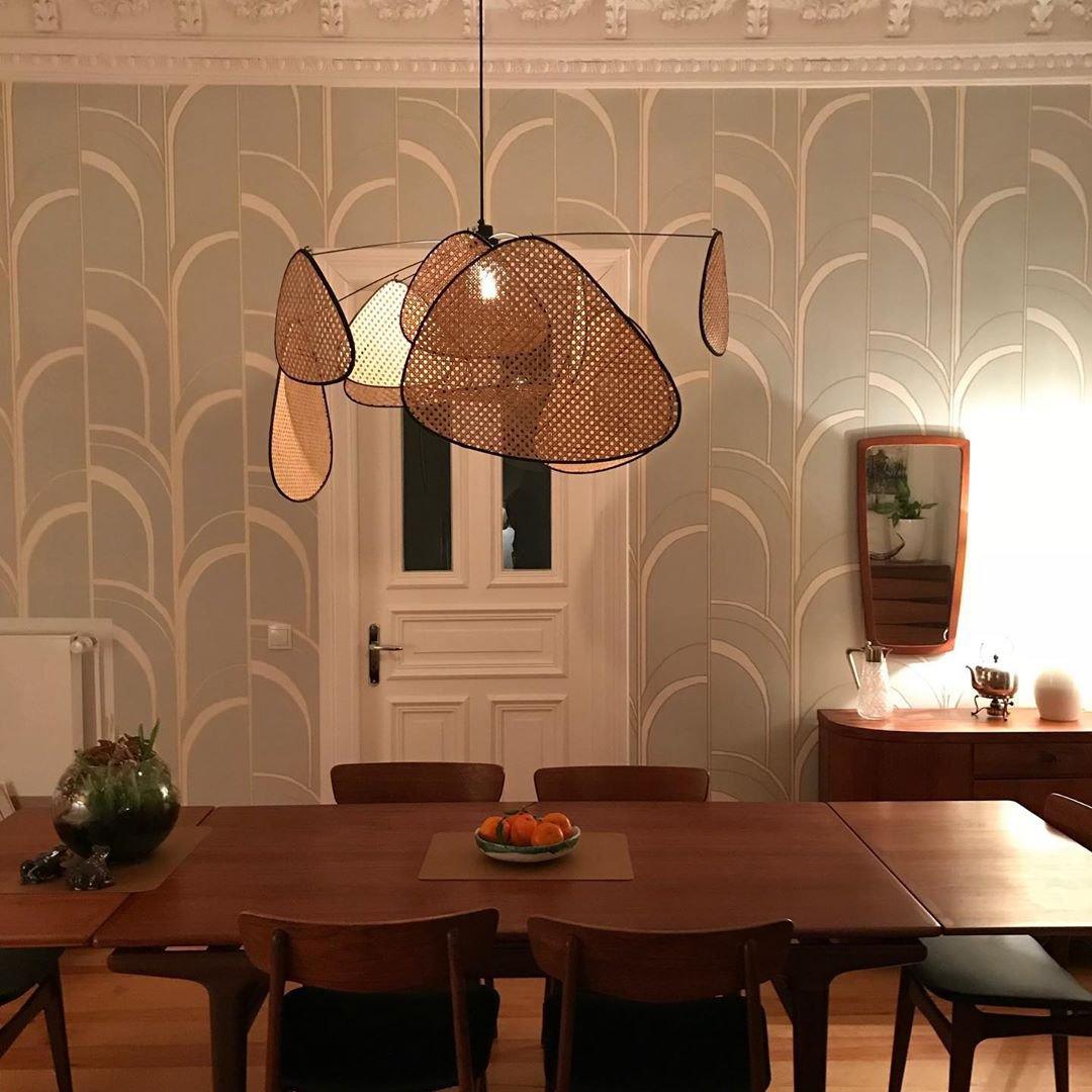 Against a patterned tan wallpaper wall, this Clérachic pendant's bamboo-weave paddles glow above a wood dining table.