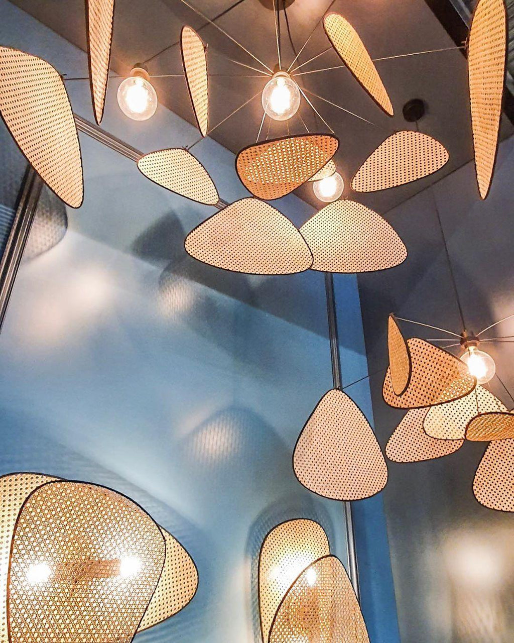 Several of this Clérachic pendant hang close together beneath a blue ceiling, their bamboo-weave paddles glowing around exposed bulbs.