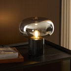 The Clérachic table lamp glows on a dark wood cabinet beside a closed book, set against a warm brown wall in a lit interior.