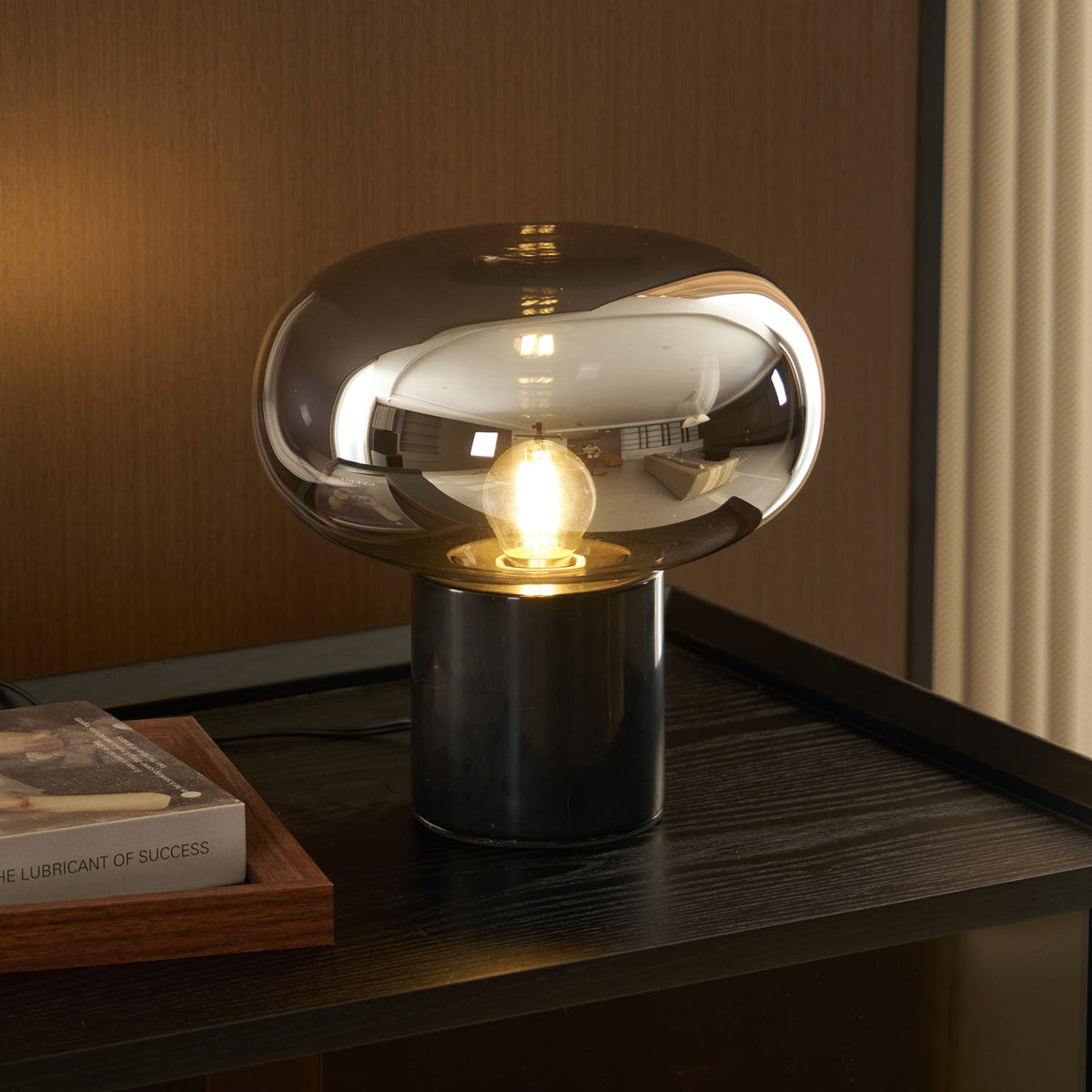 A further angle of the lit Clérachic table lamp on dark wood, its cord curving forward past the book resting beside it.