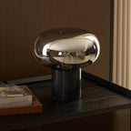 The Clérachic table lamp lit on a dark surface, its cord trailing behind the glass dome beside the propped book Evero.