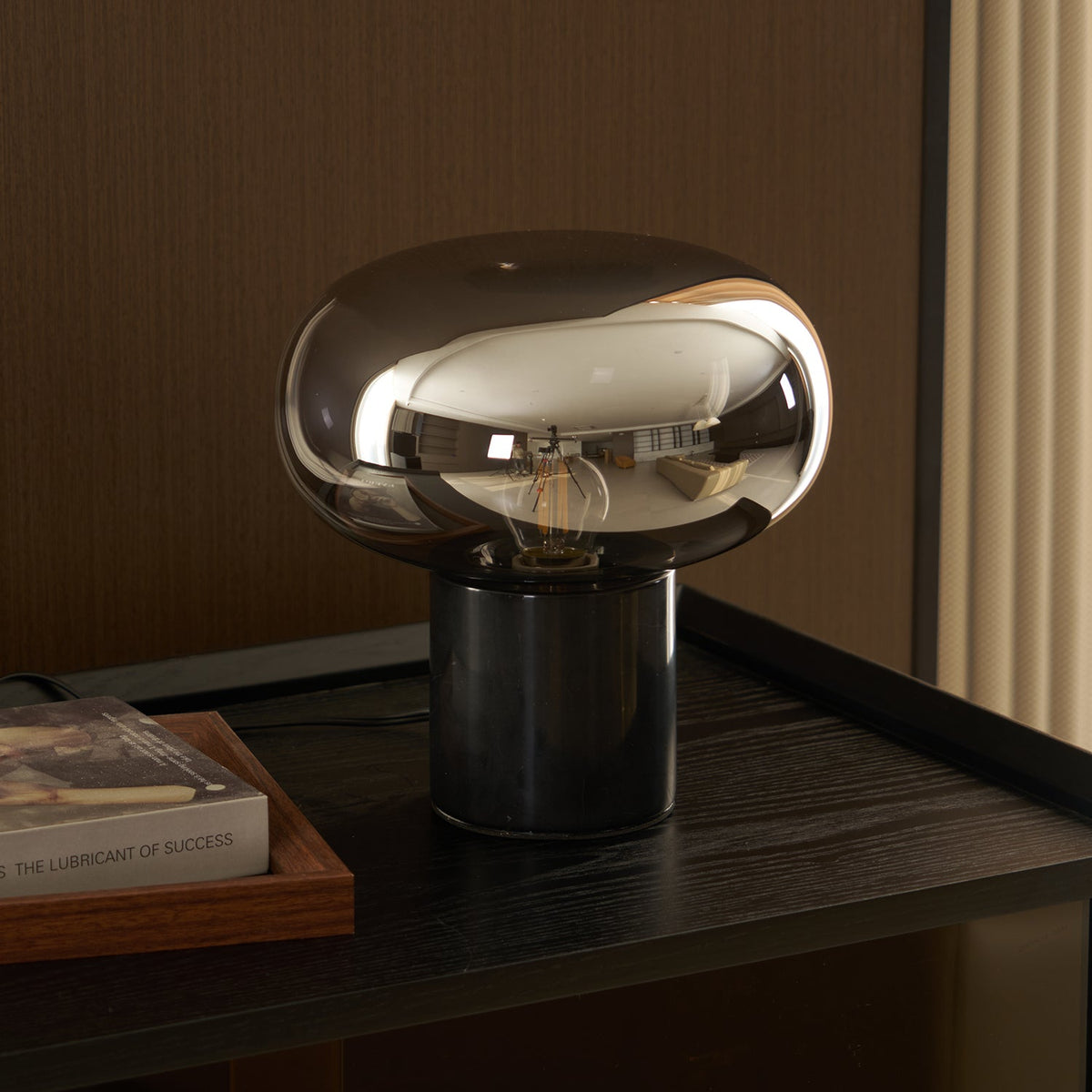 The Clérachic table lamp lit on a dark surface, its cord trailing behind the glass dome beside the propped book Evero.