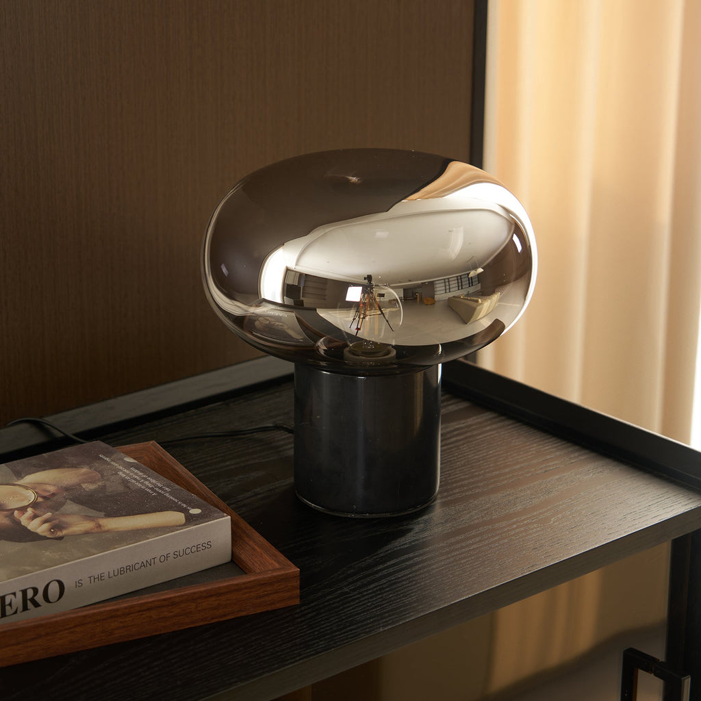Close-up of the lit Clérachic table lamp beside the book Evero, the bulb's glow visible through the glass dome.
