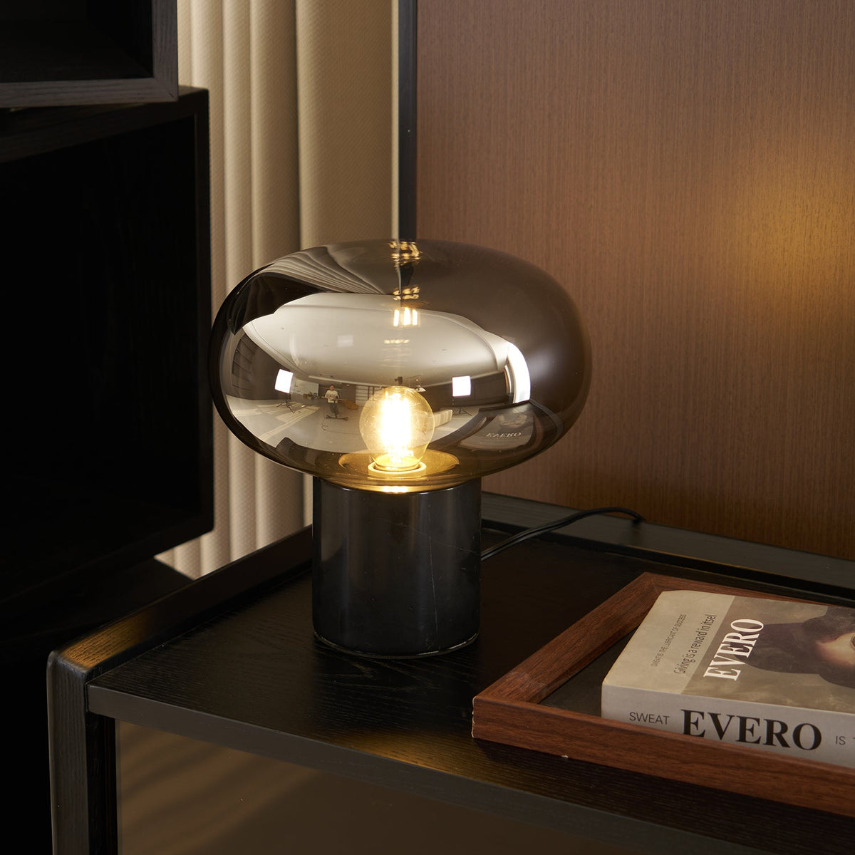 The lit Clérachic table lamp stands on a dark cabinet beside a framed book titled Evero, its glass dome glowing warmly.