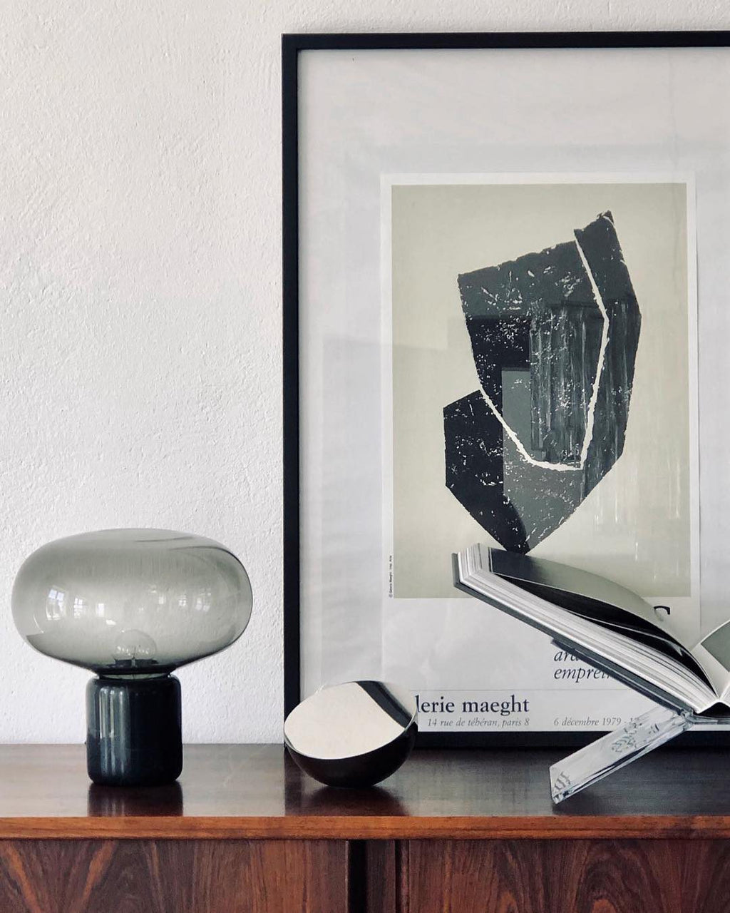 An unlit Clérachic table lamp on a dark wood sideboard, beside a framed abstract print and a stack of books.