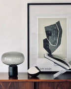 An unlit Clérachic table lamp on a dark wood sideboard, beside a framed abstract print and a stack of books.
