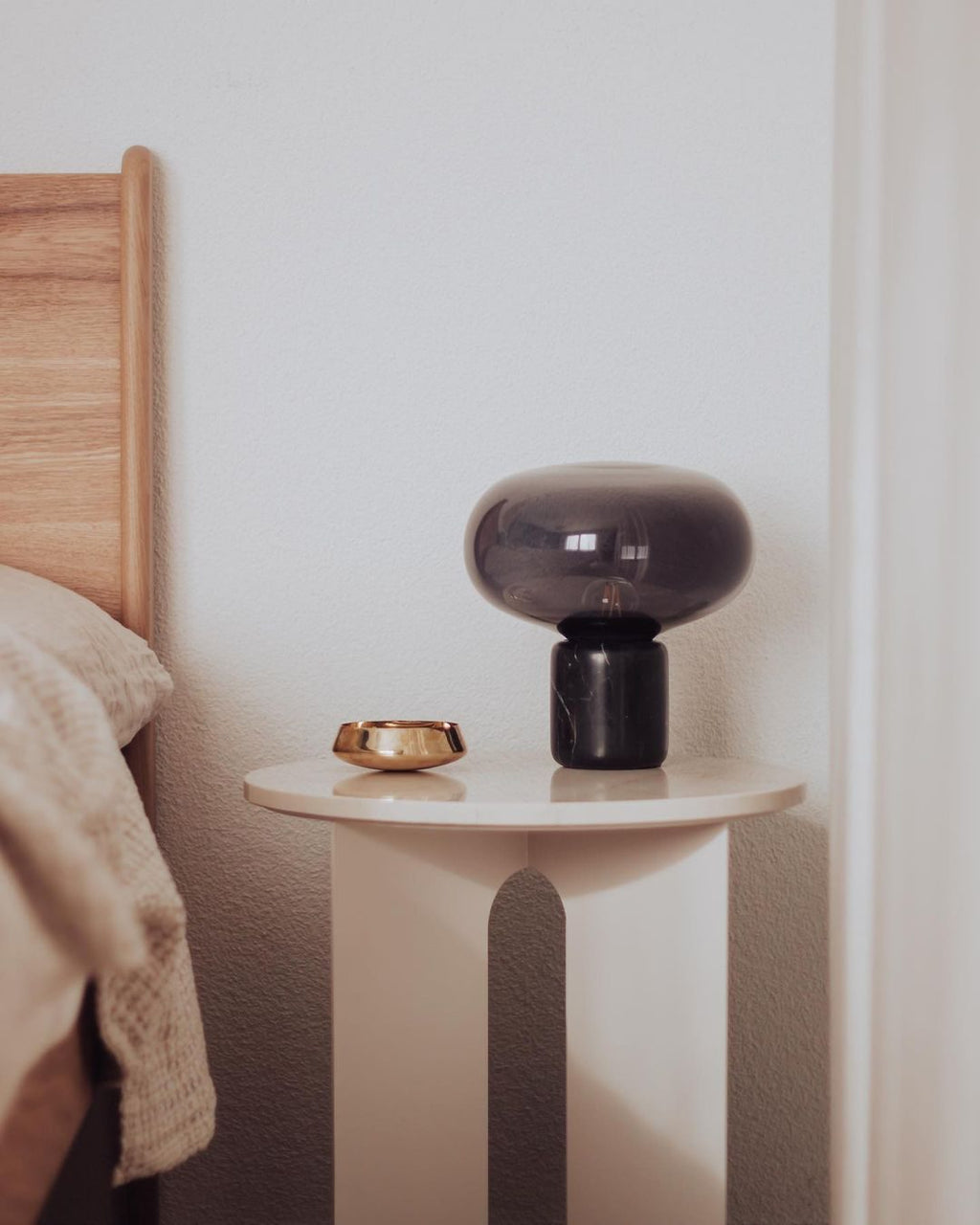 A closer crop of the Clérachic table lamp on the bedside table, with cushions and a wooden headboard behind it.