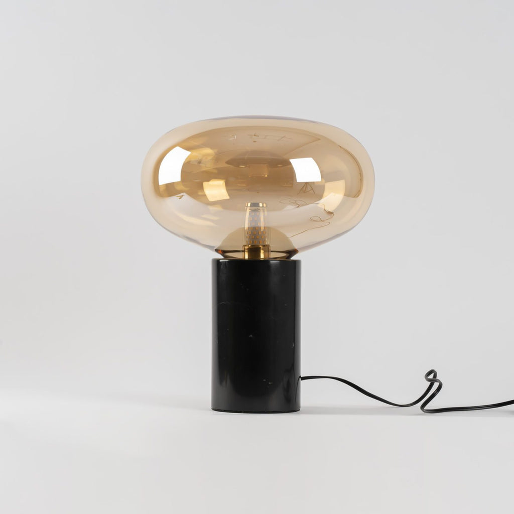 A studio shot of the amber-glass Clérachic table lamp on white, its cord looped beneath the marble base.