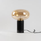A studio shot of the amber-glass Clérachic table lamp on white, its cord looped beneath the marble base.