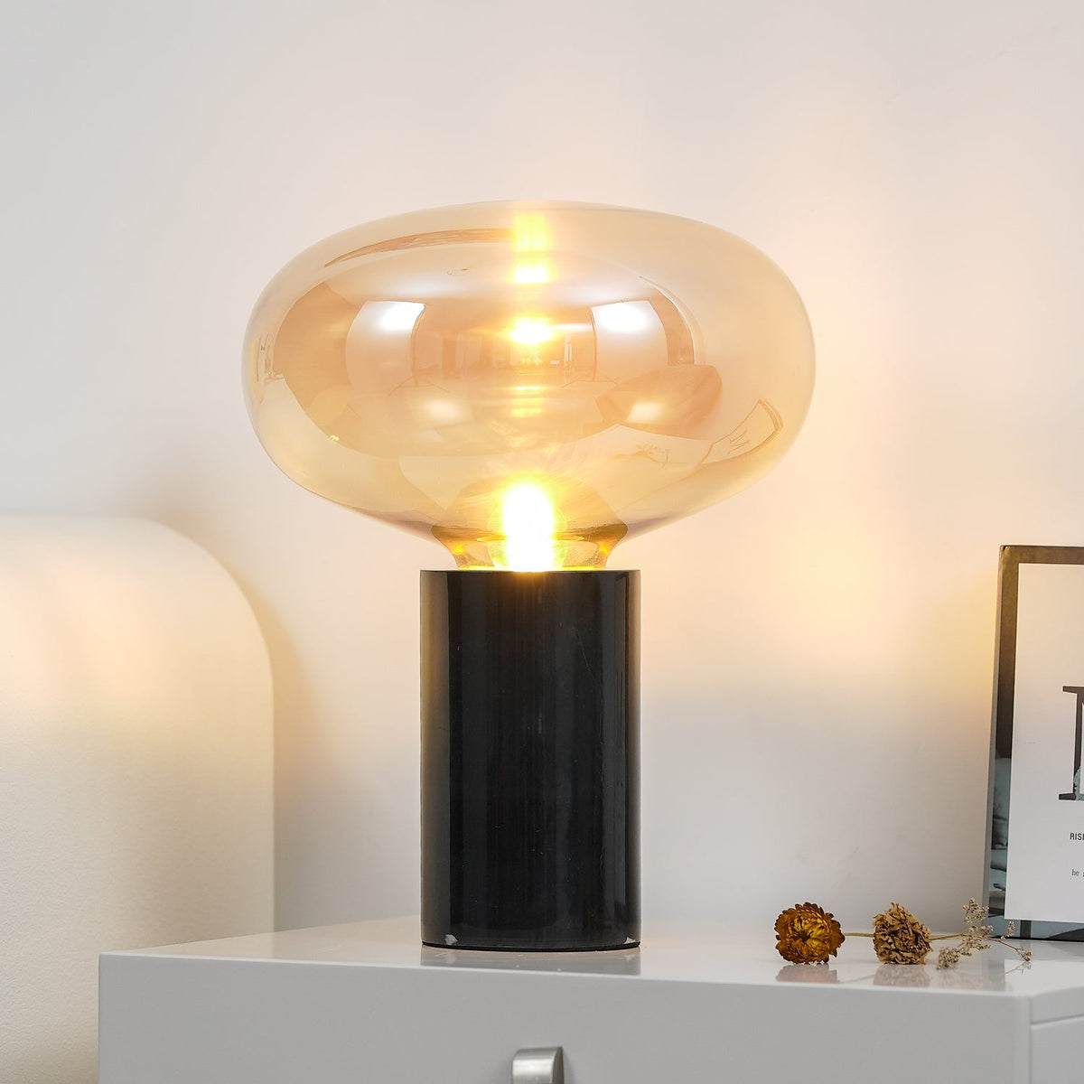 The lit amber-glass Clérachic table lamp on a white nightstand, dried flowers and a picture frame set beside it.