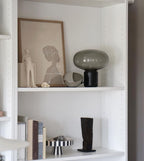 The Clérachic table lamp on a white shelf beside a small figurine, ceramic bowls and a stack of books.