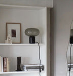 An unlit Clérachic table lamp on a white bookshelf, set between a framed portrait, a small figurine and stacked books.